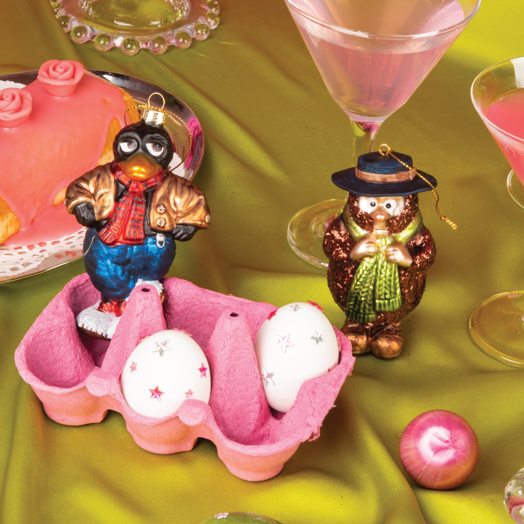 Julekuler fra HEIM Studio inspirert av Kjell Aukrusts Flåklypa – håndmalte glassfigurer av Solan med rød vest og jakke, og Ludvig med hatt og grønt skjerf. Plassert i et fargerikt bordmiljø med kake, egg og cocktailglass.