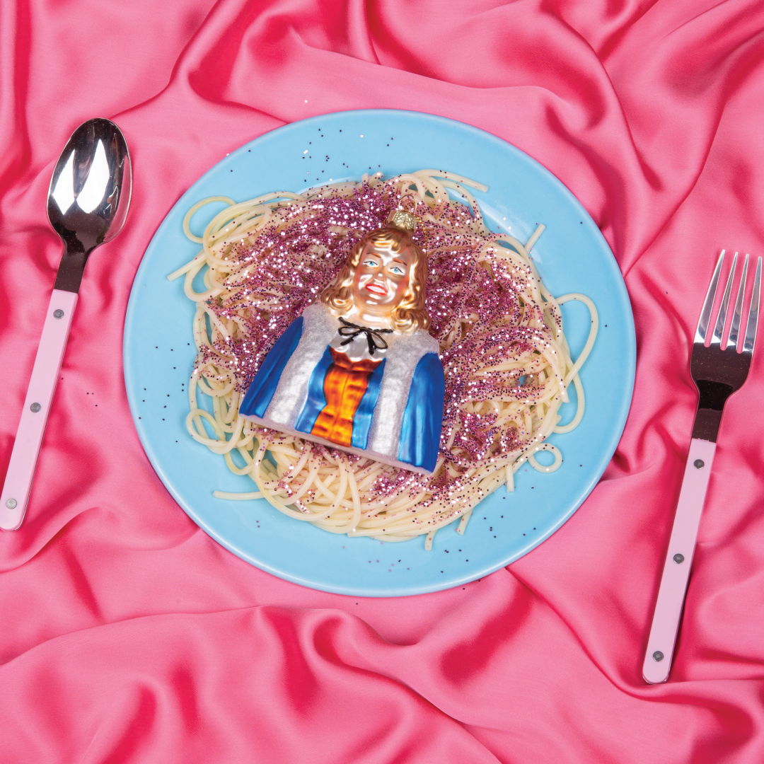 Julestjerna julekule fra HEIM Studio – håndmalt glassfigur med blondt hår, blå kappe og rød kjole. Inspirert av Hanne Kroghs ikoniske karakter. Plassert på blå tallerken med rosa bakgrunn.