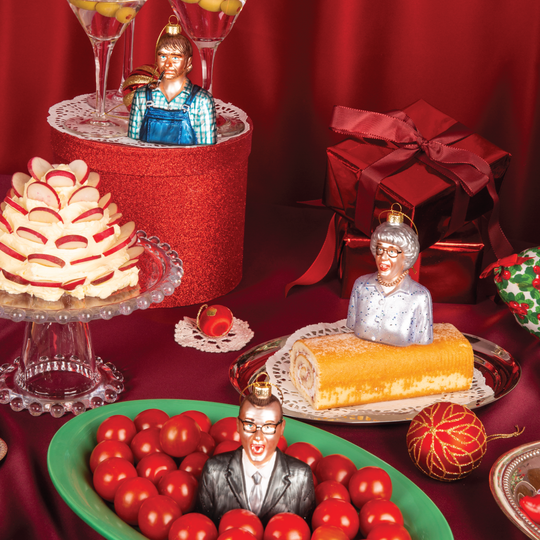 Julekuler i glass fra HEIM Studio inspirert av The Julekalender – Olaf Sand med pipe, Gjertrud Sand med perlekjede og Benny/Nåsåen i dress. Håndmalte figurer plassert i et julepyntet bordmiljø.