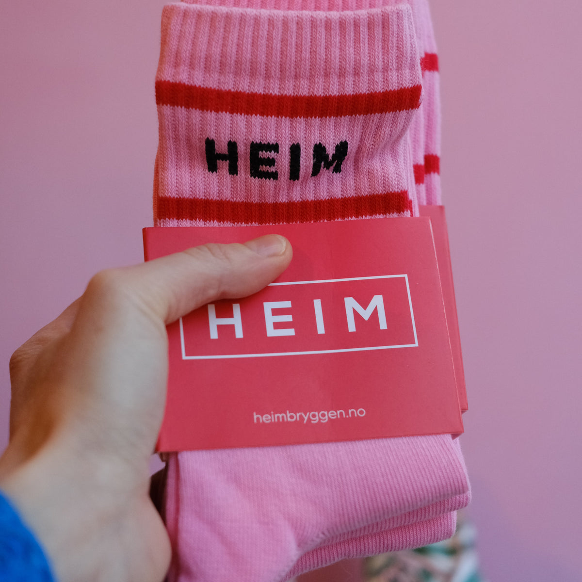 Hånd holder rosa sokker med rød og svart HEIM-logo og merke med hvit tekst på rosa bakgrunn