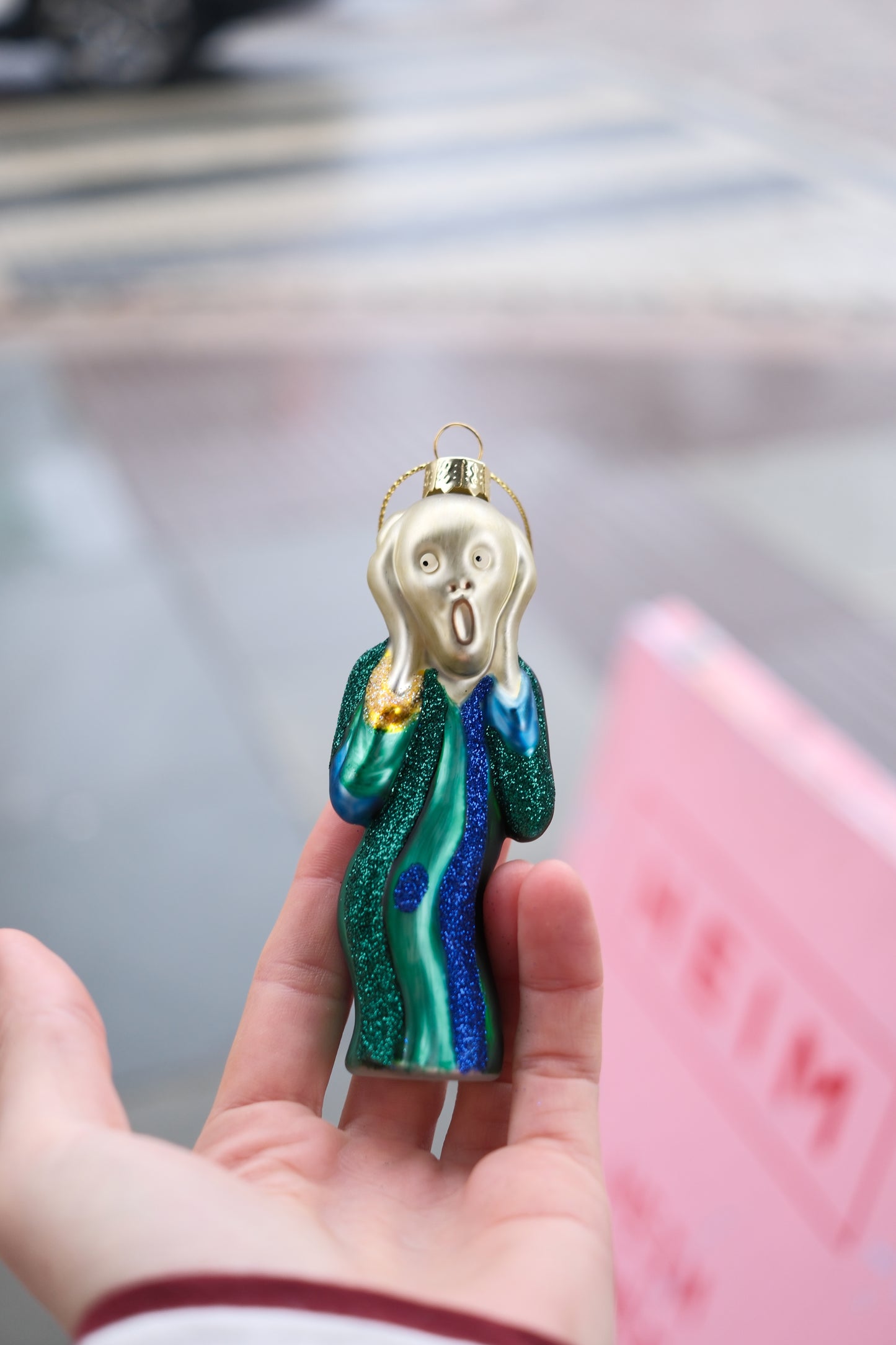 Hånd som holder julepynt formet som figur med opprørt ansikt og glittergrønn kappe mot uskarp gatebakgrunn