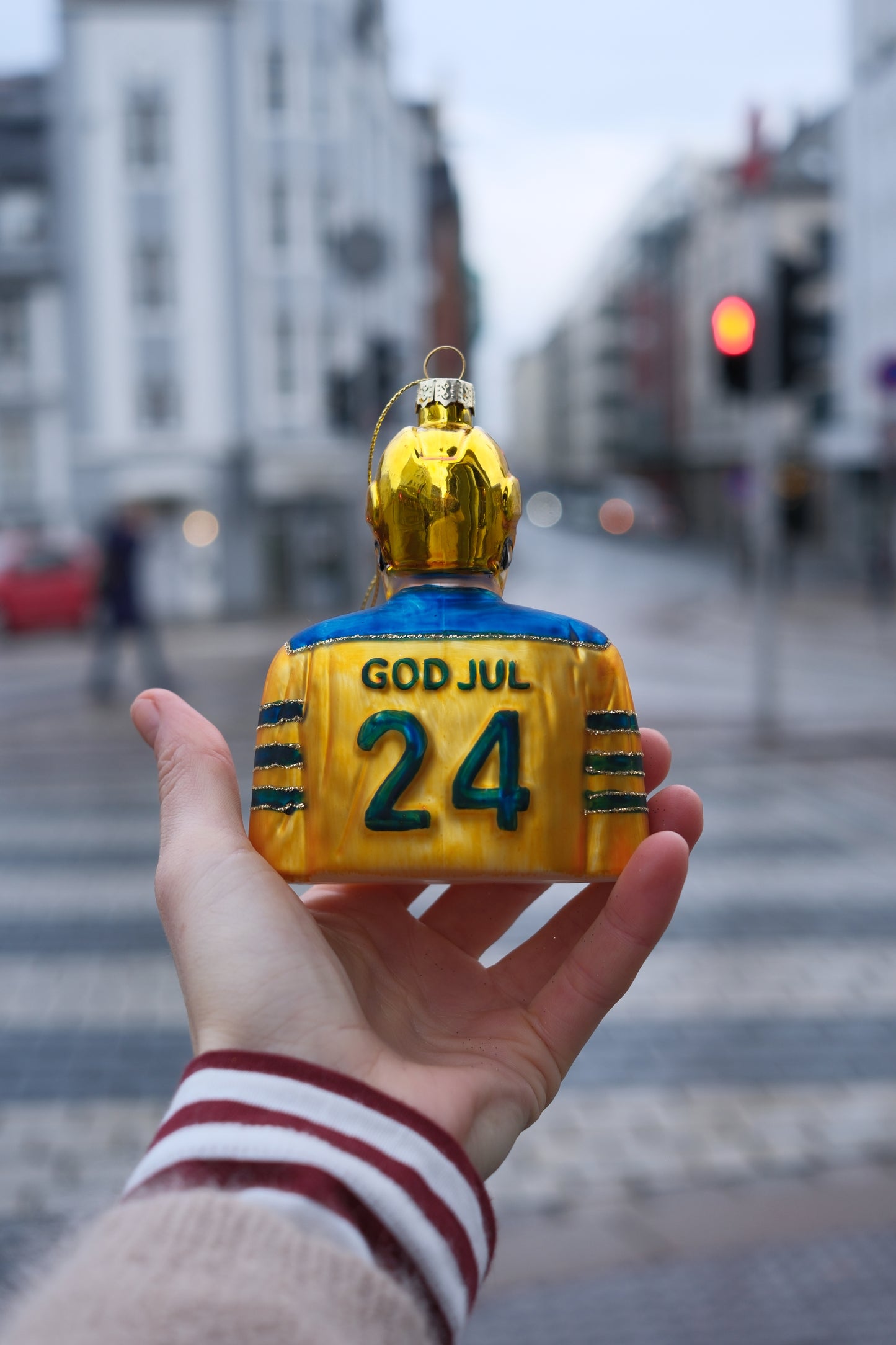 Hånd som løfter julepynt formet som figur med gullhjelm og gul blått antrekk med tekst GOD JUL 24 mot bybakgrunn