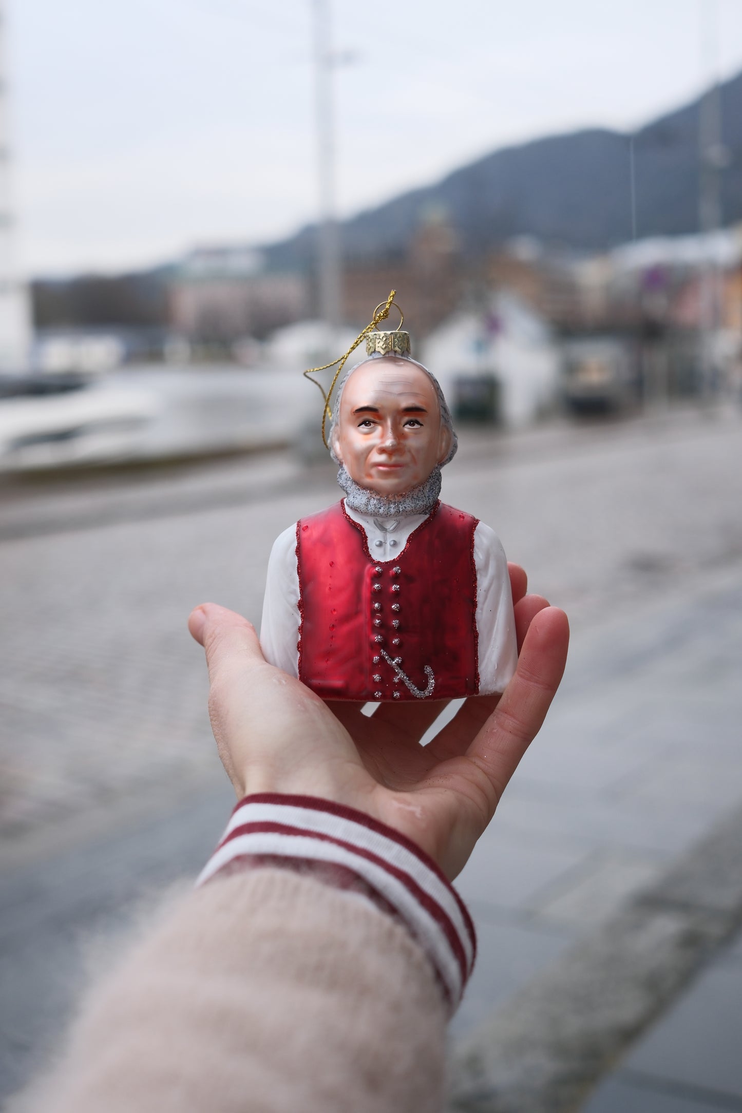 Hånd som holder julepynt formet som mann i rød vest og hvit skjorte med sløret bymiljø i bakgrunnen