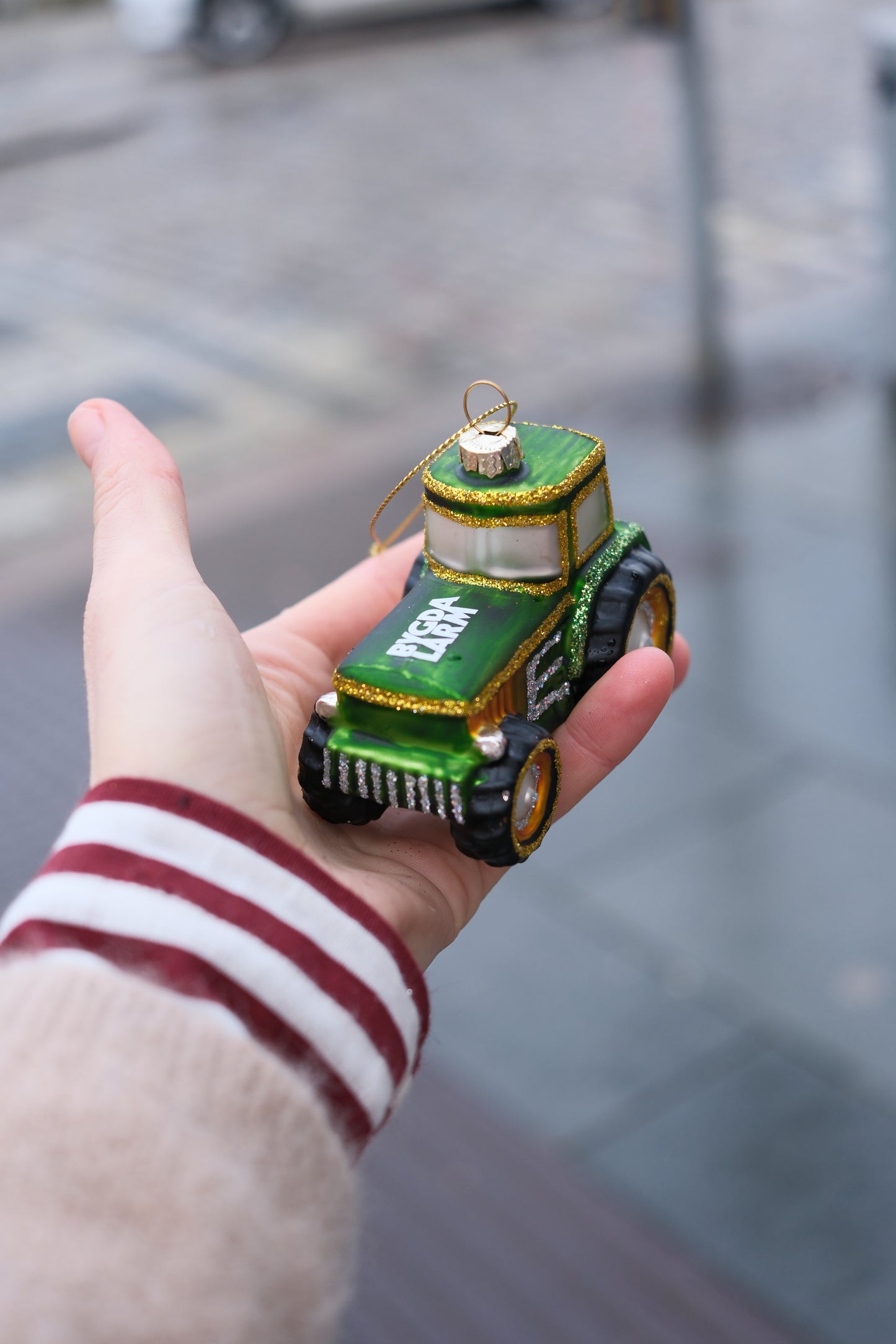 Hånd som holder grønn julepynt formet som traktor med gullglitter mot uskarp gatebakgrunn
