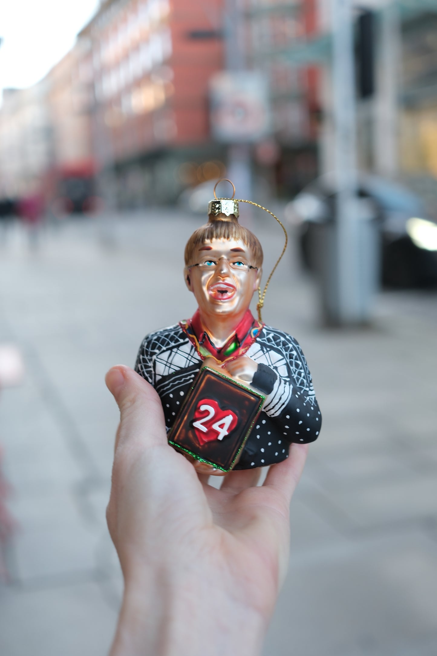 Hånd som holder julepynt formet som person med briller og mønstret genser med tall 24 på bok foran bymiljø