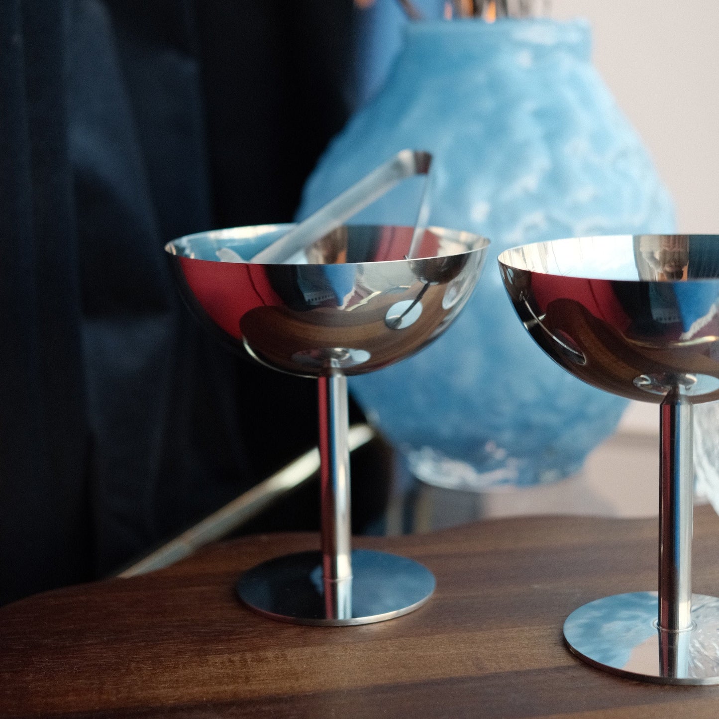 Cocktail glass i stål - by Heim design med blank rustfri overflate og elegant rund form på trebord foran blå vase