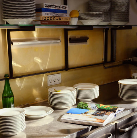 Stablede hvite tallerkener i ulike størrelser på rustfritt bord med bok og grønn flaske under hylle med oppbevaring