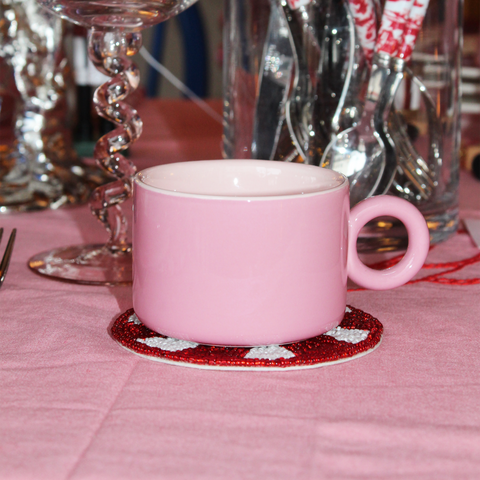 Rosa keramisk kopp med rundt glassunderlag på rosa duk i omgivelser med glass og bestikk bakgrunn