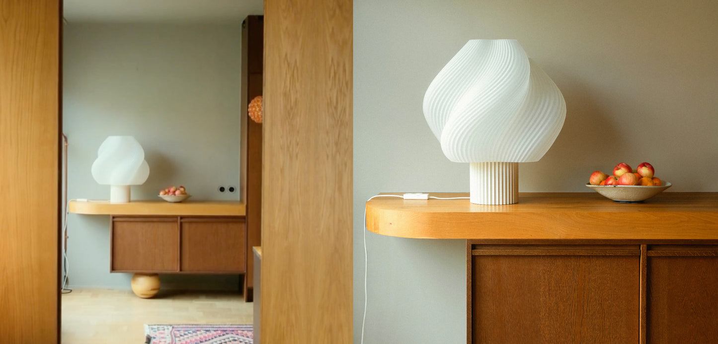 Hvit bordlampe med bølget design plassert på treskap med skål fylt med epler ved vegg i rom med speil