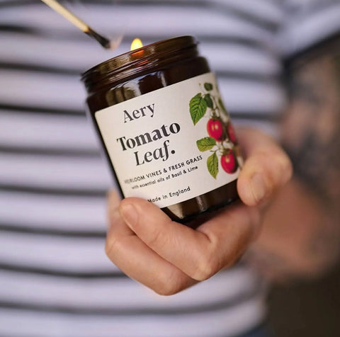 Hånd som holder brunt glasslys med etikett og illustrasjon av tomatplante mot bakgrunn med stripete klær