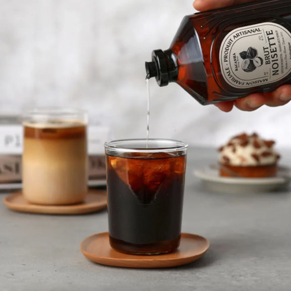 Cinnamon Organic Syrup - By Bacanha helles over isbiter i et enkelt glass med kaffe og kake i bakgrunnen