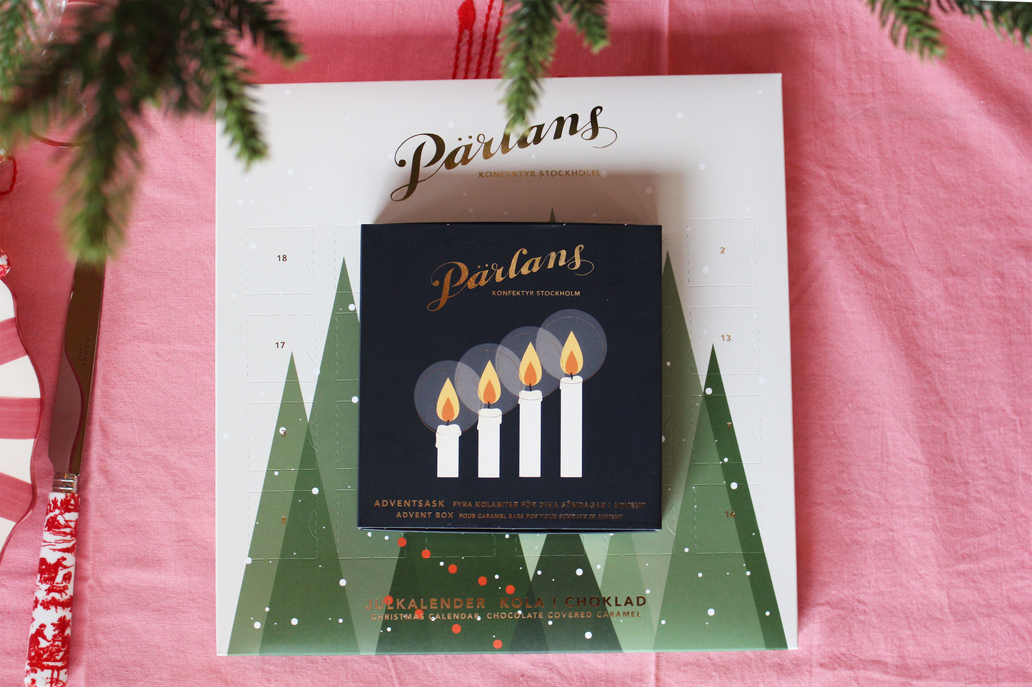 Adventskalender fra Pärlans Konfektyr med fire telys og grantrær på rosa bakgrunn for vinterstemning