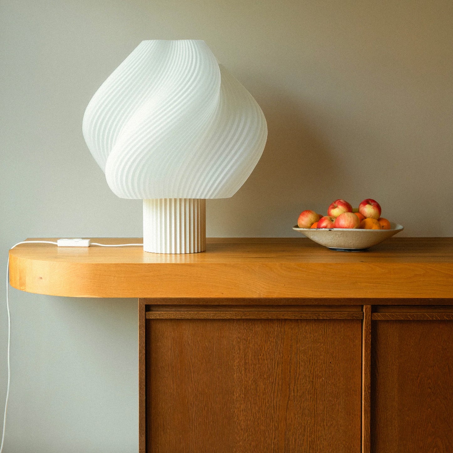 Hvit bordlampe med bølgende form på lyst trebord ved treskap og skål med røde epler mot beige vegg