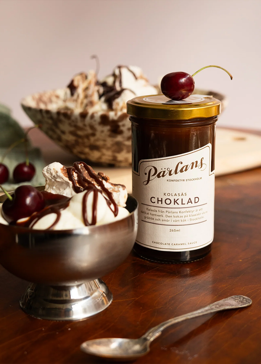 Glass med sjokoladesaus med kirsebær på lokk ved siden av dessertskål med krem og sjokoladesaus på trebord