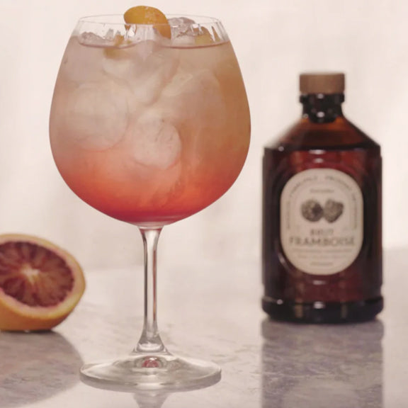 Raspberry Organic Syrup - By Bacanha servert i glass med is og sitrus, flaske i bakgrunnen på lys overflate