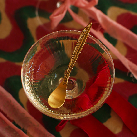 Stella Liten Skje - By Doing Goods i gullfarget metall formet som en fjær i glass på fargerik bakgrunn.