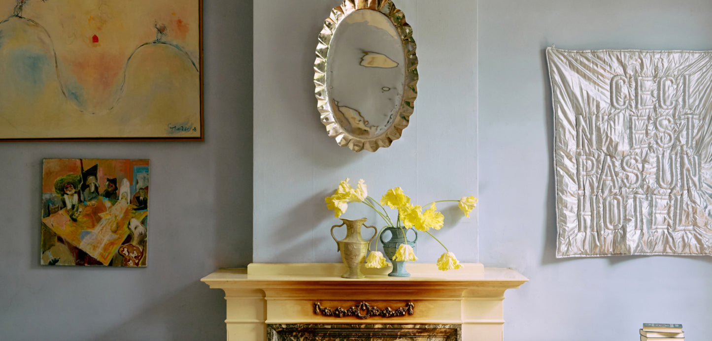 Interiørscene med peis, ovalt speil, bukett med gule blomster i dekorative vaser og kunstverk på veggen